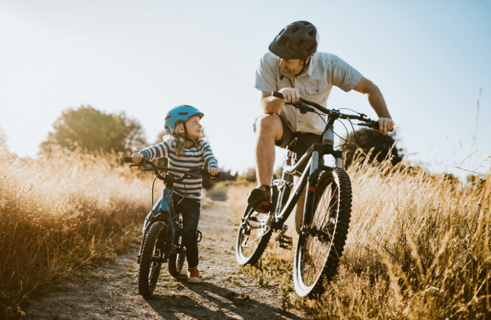 Man and child riding bikes together on a dirt path through a grassy area.